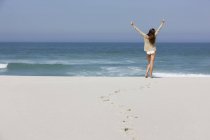 Vista trasera de la mujer feliz de pie en la playa de arena con los brazos levantados - foto de stock