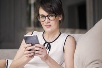 Вдумчивая женщина в очках держит смартфон и смотрит на диван — стоковое фото