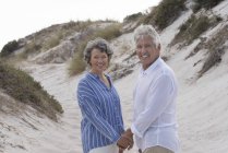 Feliz pareja de ancianos de pie en la playa de arena de la mano y mirando a la cámara - foto de stock