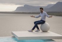 Человек, сидящий на каменном шаре и жестикулирующий на берегу озера — стоковое фото