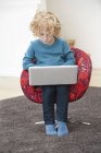 Menino bonito com cabelo loiro usando um laptop em poltrona em casa — Fotografia de Stock