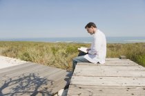 Hombre sentado en el paseo marítimo en la naturaleza y leyendo un libro - foto de stock