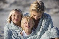 Портрет усміхненої матері і дітей, загорнуті в хустку, що сидить на пляжі — стокове фото