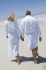 Visão traseira do casal em roupões de banho andando na praia de areia de mãos dadas — Fotografia de Stock