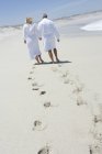 Rückansicht eines Paares in Bademänteln, das Händchen haltend am Sandstrand spaziert — Stockfoto