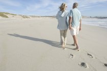 Rückansicht eines reifen Paares, das Händchen haltend am Strand spaziert — Stockfoto