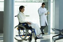 Paziente femminile seduta in sedia a rotelle e medico di sesso maschile in piedi su sfondo in ospedale — Foto stock