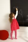 Little girl in red dress drawing on a blackboard in classroom — Stock Photo