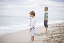 Мальчики стоят на песчаном пляже в пасмурной тени и смотрят на вид — стоковое фото