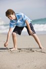 Teenage boy drawing in sand on sunny beach — Stock Photo