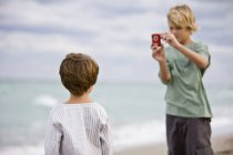 Хлопчик фотографує брата з цифровою камерою на пляжі — стокове фото