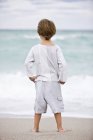Вид сзади на маленького мальчика, стоящего на пляже и смотрящего на море — стоковое фото