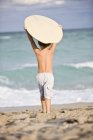 Rear view of little boy holding body board over head on sandy beach — Stock Photo