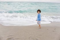 Мальчик с кудрявыми волосами стоит на пляже и смотрит в сторону — стоковое фото