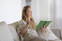 Mujer enfocada leer libro y comer galletas en el sofá en casa - foto de stock