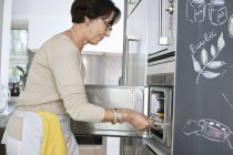 Senior woman putting tray of seafood into oven in kitchen — Stock Photo