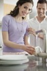 Женщина стирает посуду на кухне с мужем на заднем плане — стоковое фото