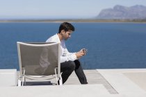 Красивий чоловік сидить на шезлонгу на березі озера і використовує смартфон — стокове фото
