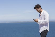 Задумчивый мужчина в белой рубашке с помощью смартфона на берегу озера — стоковое фото