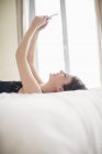 Жінка сміється лежить на ліжку і використовує мобільний телефон — стокове фото