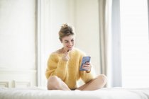 Frau im gelben flauschigen Pullover telefoniert und isst Schokolade auf dem Bett — Stockfoto