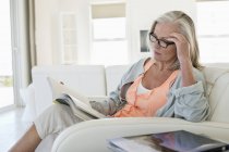 Старша жінка сидить на дивані і читає книгу вдома — стокове фото