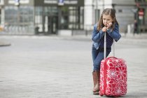 Grimacing little girl standing with luggage on street — Stock Photo