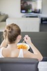 Дівчина їсть макарони сидячи на дивані і дивиться телевізор вдома — стокове фото
