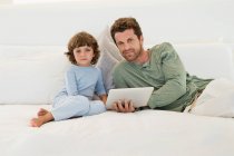 Чоловік тримає цифровий планшет, сидячи на ліжку зі своїм сином — стокове фото