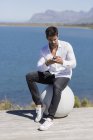 Homem confiante sentado na bola de pedra e usando smartphone na margem do lago — Fotografia de Stock