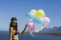 Stylish young woman holding colorful balloons on lake shore against blue sky — Stock Photo