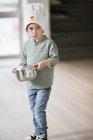 Мальчик в шляпе шеф-повара несет кастрюлю и смотрит в сторону — стоковое фото