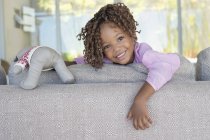 Portrait of smiling little girl holding teddy bear on sofa in room — Stock Photo
