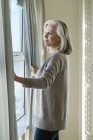 Femme âgée réfléchie debout à la fenêtre à la maison — Photo de stock