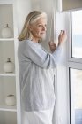 Senior woman looking through window at home — Stock Photo