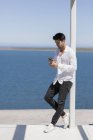 Hombre seguro de sí mismo apoyado en el poste en la orilla del lago y el uso de smartphone - foto de stock