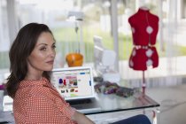 Durchdachte Modedesignerin sitzt mit Laptop im Hintergrund im Büro — Stockfoto