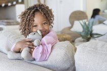Retrato de niña sonriente abrazando oso de peluche en el sofá en la habitación - foto de stock
