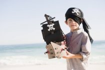 Мальчик в пиратском костюме играет с игрушечной лодкой на пляже — стоковое фото