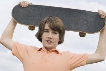 Portrait of teenage boy holding skateboard over head — Stock Photo