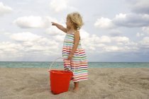 Linda niña sosteniendo un cubo de arena en la playa y apuntando al mar - foto de stock