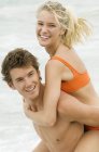 Cheerful man giving woman piggyback ride on beach — Stock Photo
