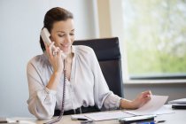 Усміхнена бізнес-леді розмовляє по стаціонарному телефону в офісі — стокове фото