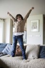 Portrait of cheerful boy standing on sofa with hands up at home — Stock Photo
