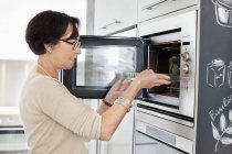 Mujer mayor poniendo bandeja en el horno - foto de stock