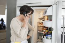 Старша жінка дивиться на холодильник і розмовляє на мобільний телефон на кухні — стокове фото