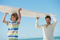 Mann und Sohn tragen Surfbrett am Strand — Stockfoto