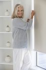 Mulher sênior ajustando cortina de janela em casa e olhando para a câmera — Fotografia de Stock