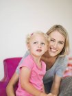 Mujer sonriente sentada con linda hija rubia en casa - foto de stock
