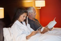Mulher usando tablet digital com marido ler livro no quarto de hotel — Fotografia de Stock